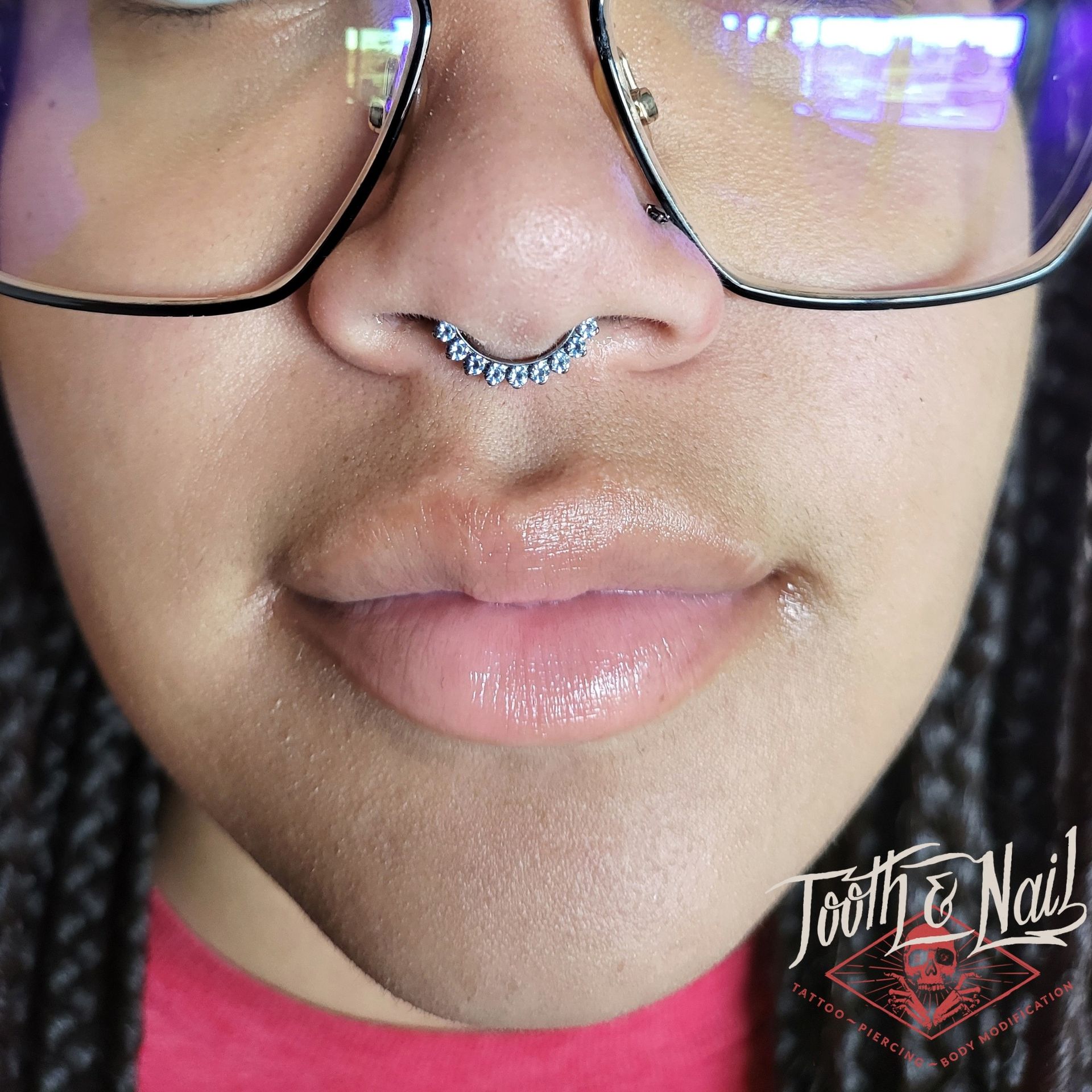 Close-up of a nose with a silver septum ring, glasses, and glossy lips with a Tooth & Nail watermark.