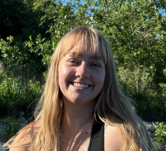Smiling person with long blonde hair standing outdoors in sunlight, with green trees behind them