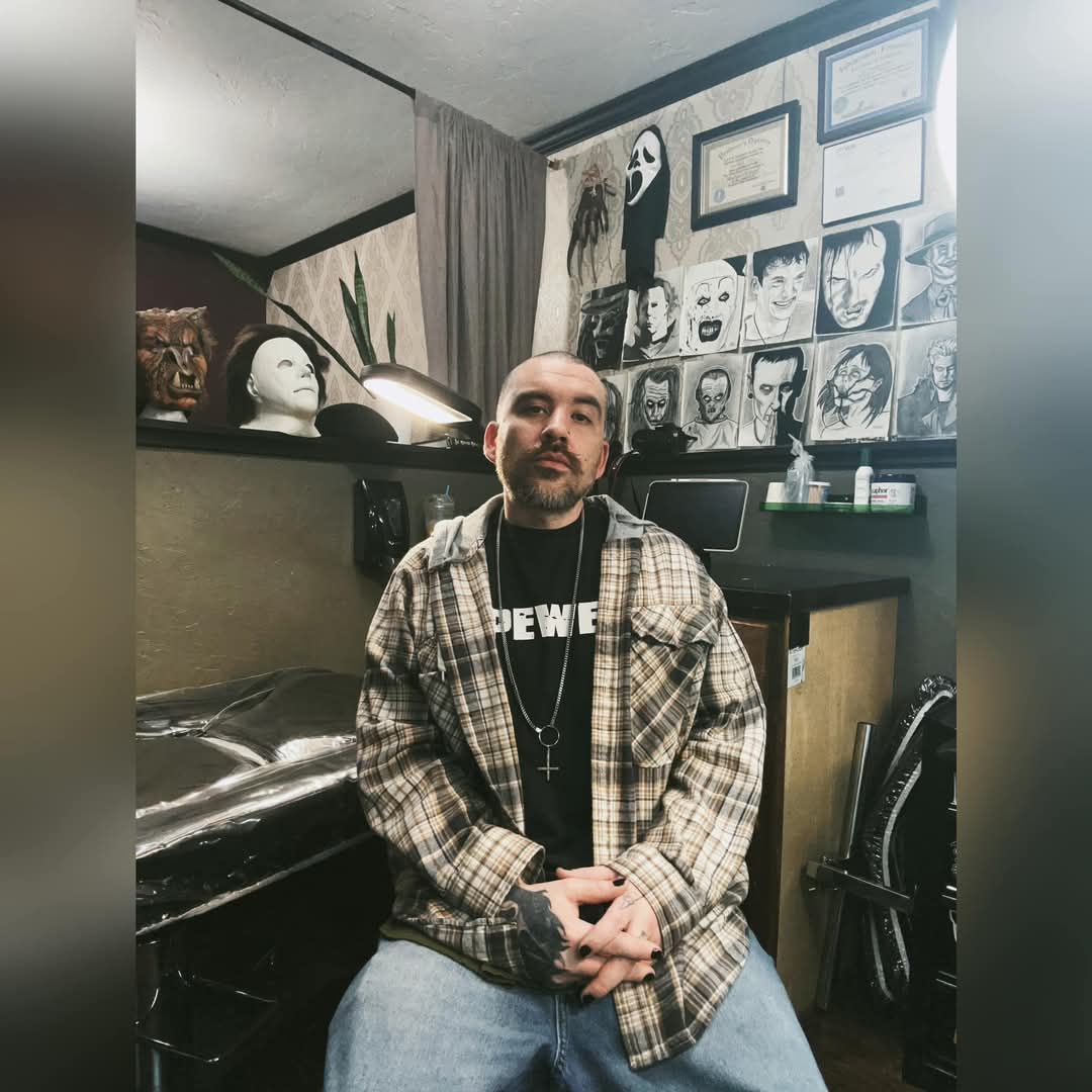 Person seated in a tattoo studio, surrounded by framed artwork and portraits on the wall.