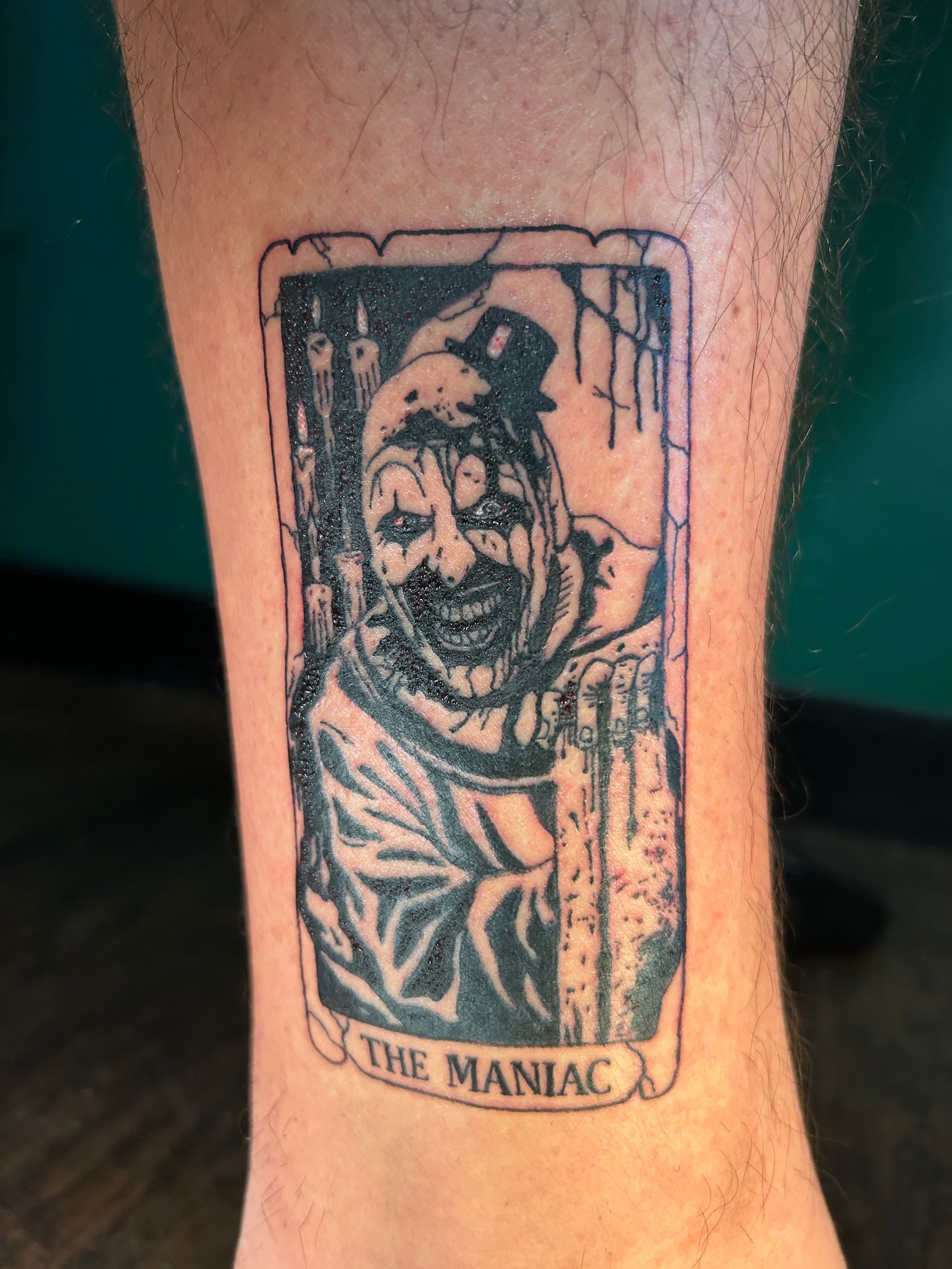 Black-and-white tattoo of a menacing clown with the words “THE MANIAC” on a forearm