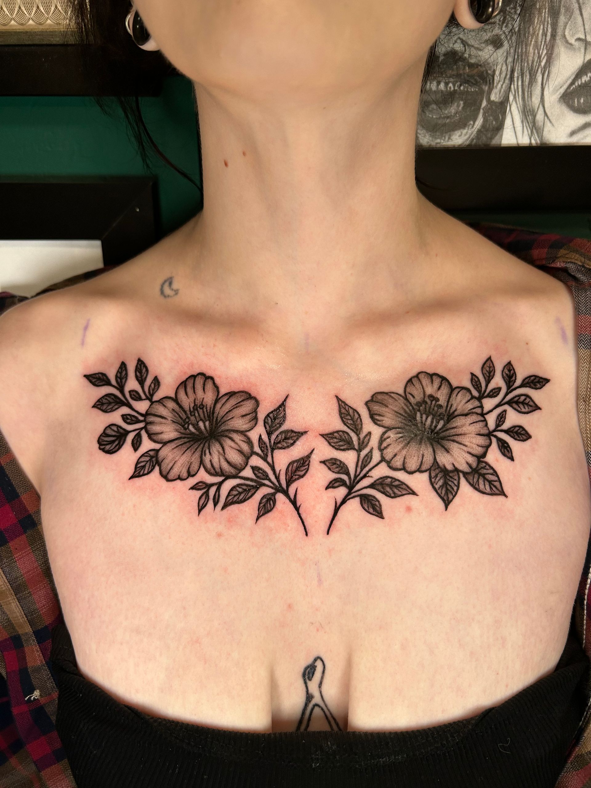 Neckline floral chest tattoo with two large black flowers and leaves on a person’s upper chest