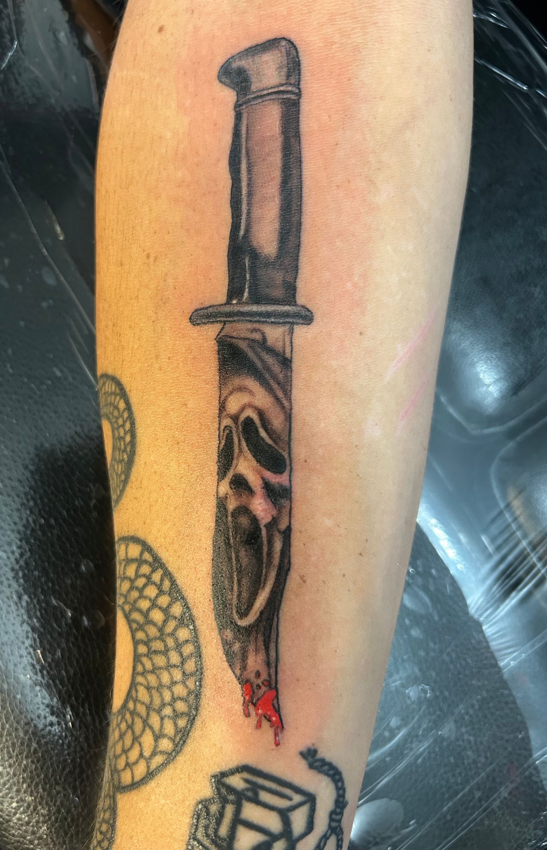 Close-up of a forearm tattoo showing a vertical dagger with a skull-like handle and red drips, beside a snake pattern