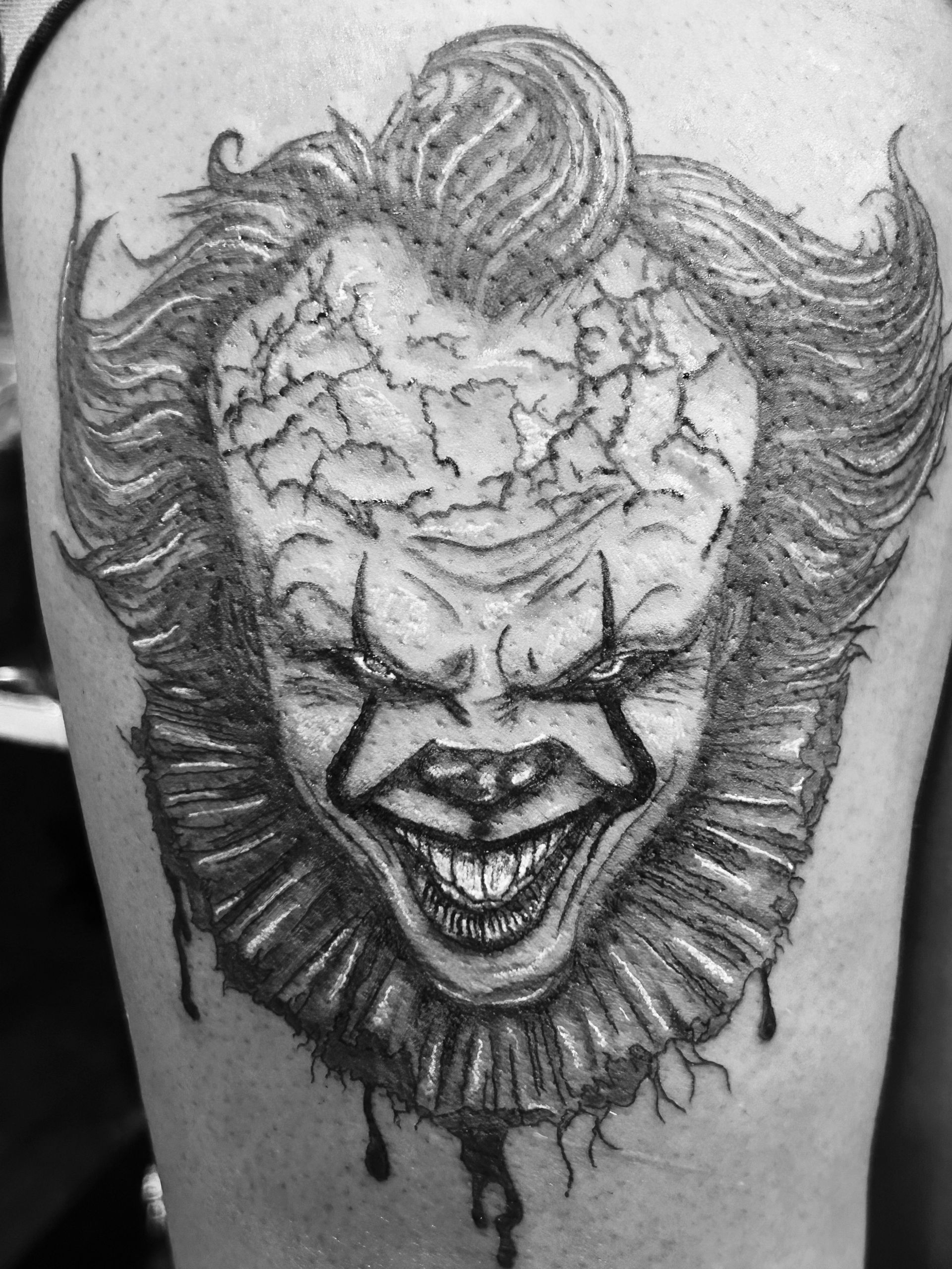 Black-and-gray clown tattoo with cracked forehead, wild hair, and dripping beard on skin