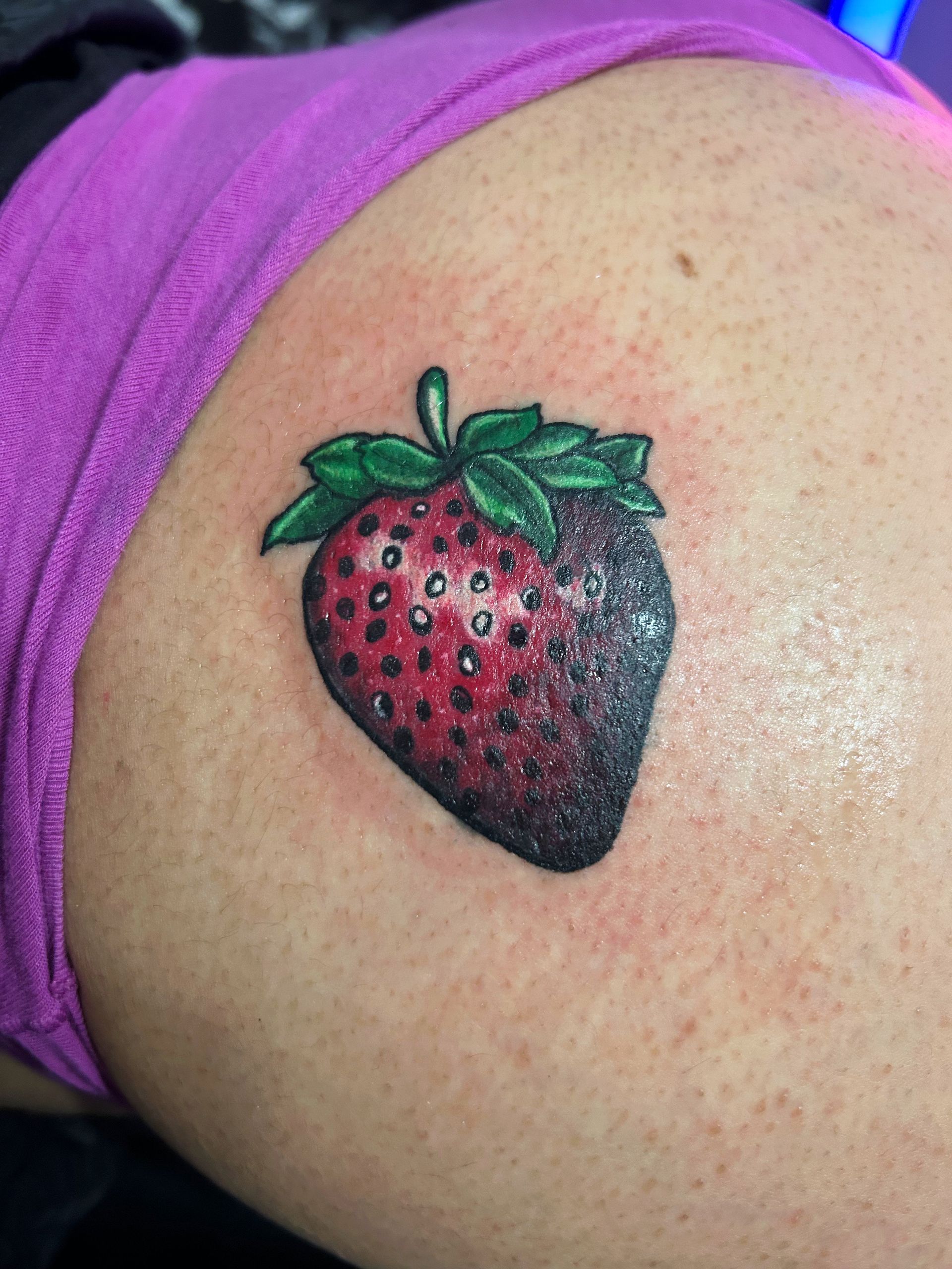 Strawberry tattoo with green leaves on a person's upper arm, beside a purple shirt strap