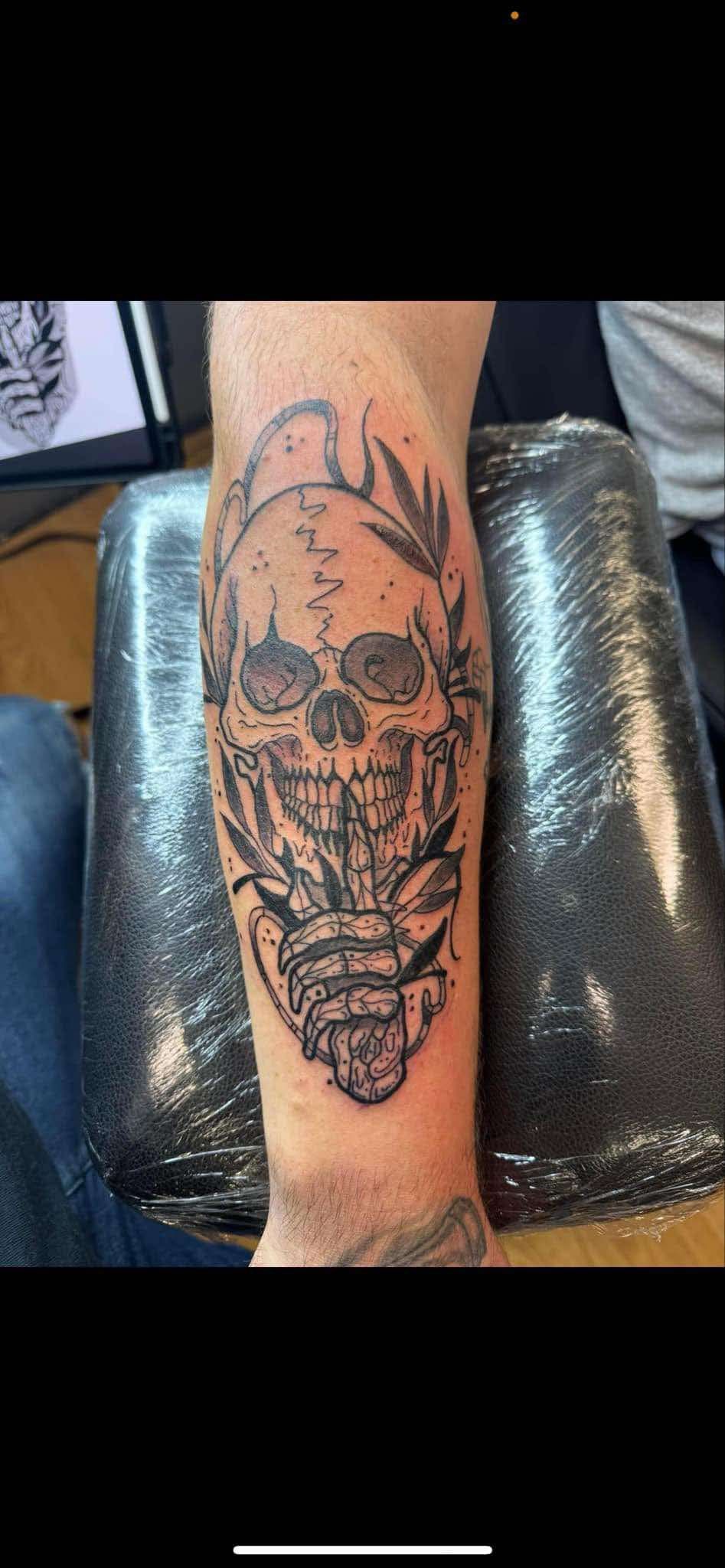 Arm tattoo of a skull-faced figure with horn-like shapes on a person’s forearm, viewed indoors on a couch.