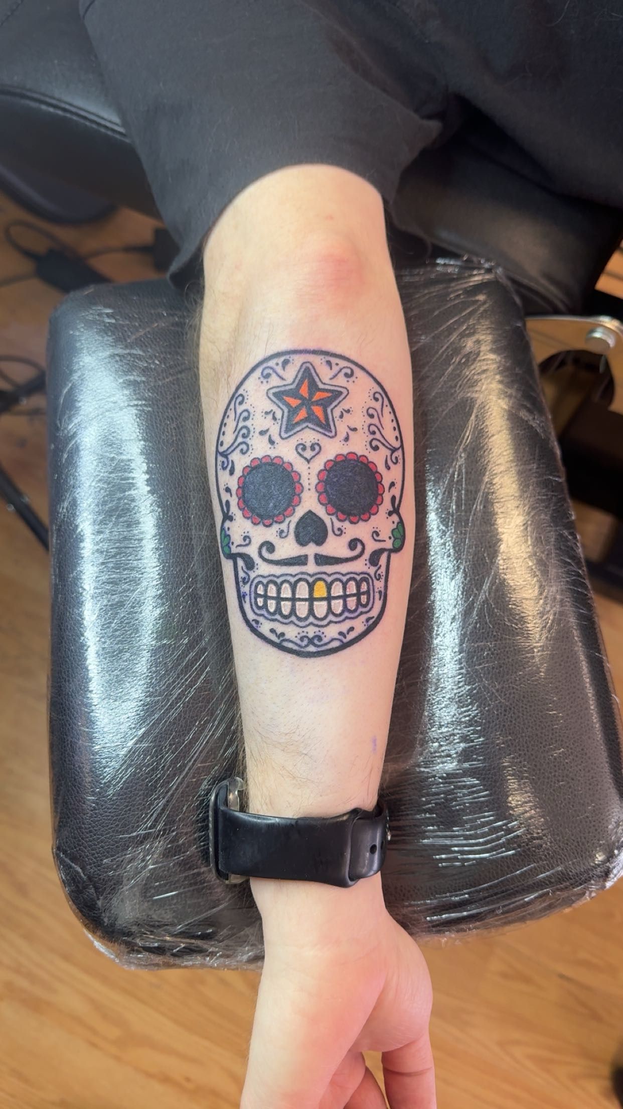 Forearm resting on a black armrest, showing a colorful sugar skull tattoo.