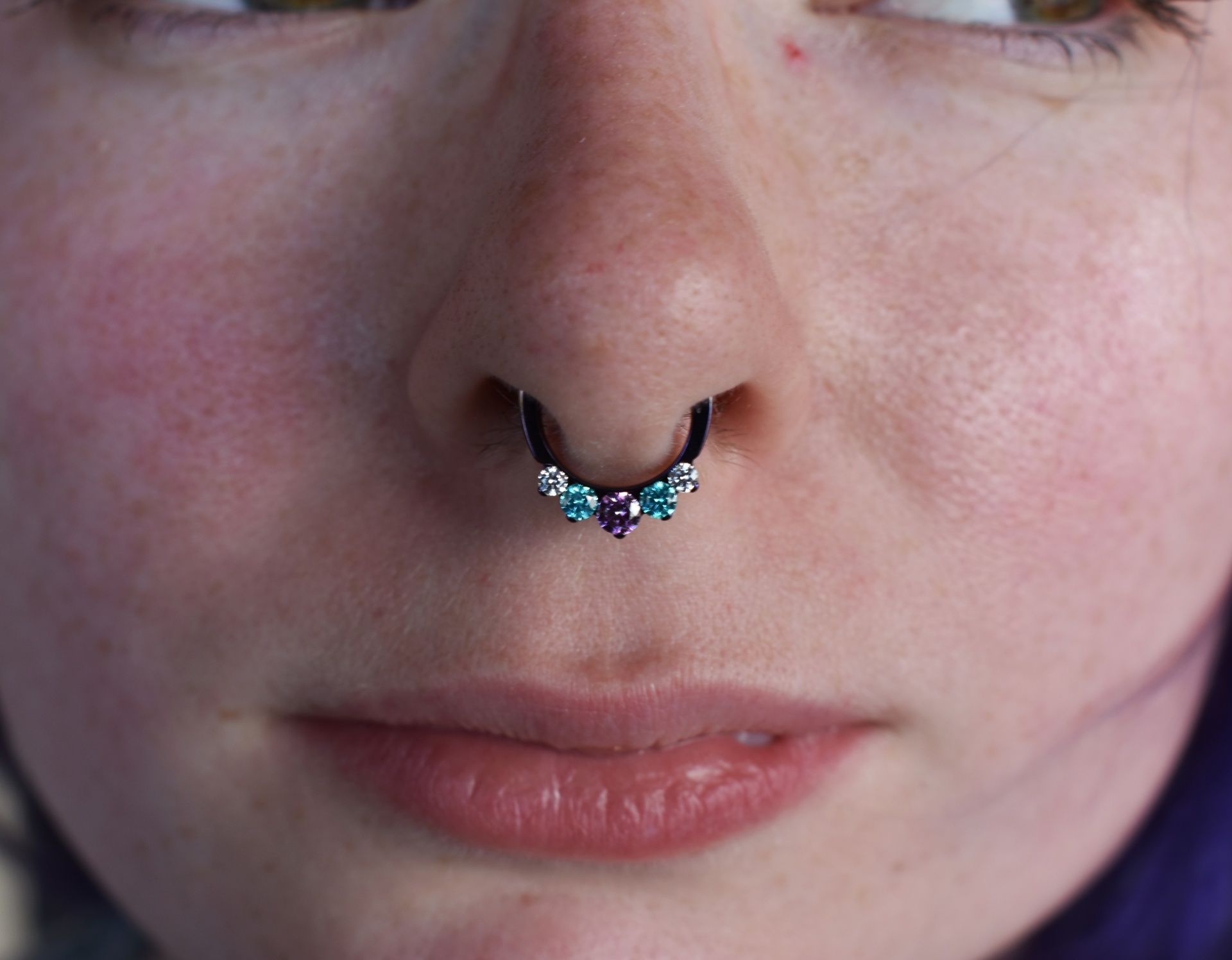 Close-up of a face with a black septum ring and teal and purple gem accents