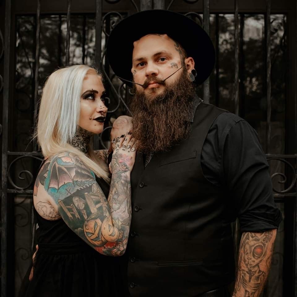 Tattooed couple posing together outdoors, the man in a black hat and the woman with platinum hair.
