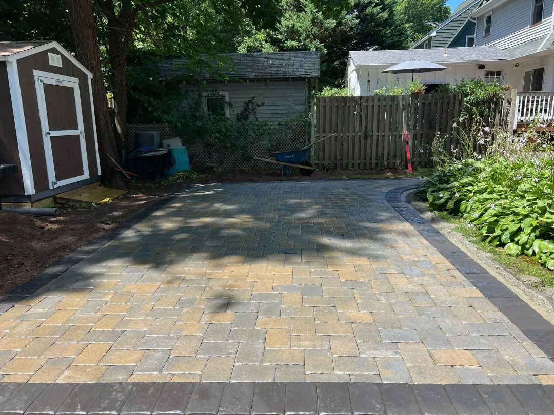 Brick paved driveway with brown and tan bricks, a brown shed, and a wooden fence.