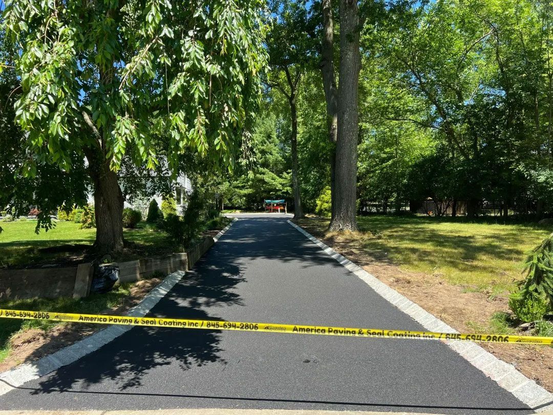 Newly paved asphalt driveway with white border, yellow caution tape, lined by trees and grass.
