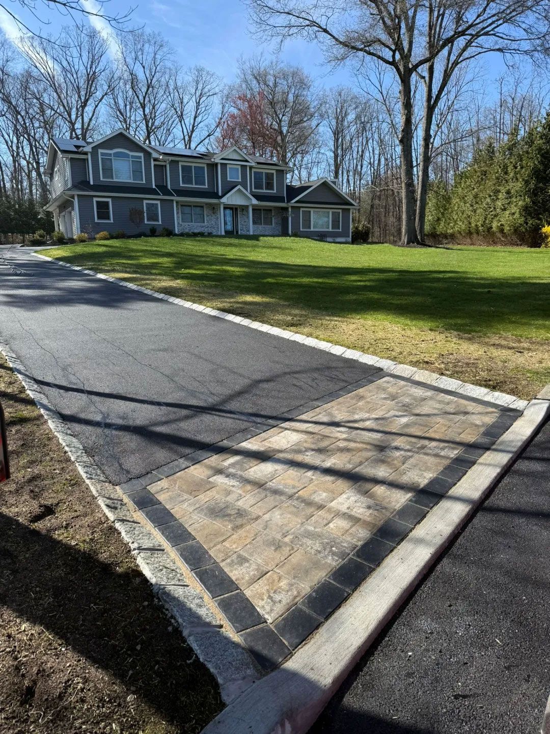 Asphalt driveway with stone pavers leading to a gray house with a green lawn and trees.