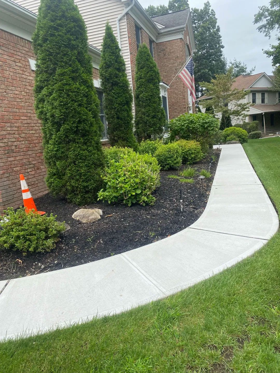 Sidewalk curves along a house with bushes and evergreen trees in a landscaped yard.