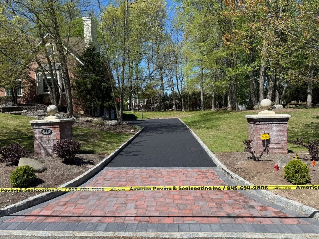 Newly paved driveway with brick border and decorative pillars.  House and trees in the background.