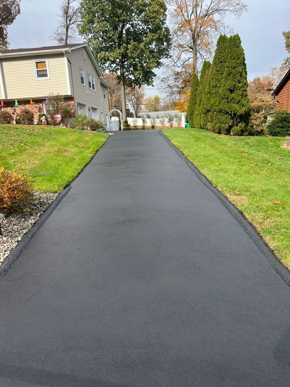 Newly paved black asphalt driveway between green lawns, leading towards a residential area.