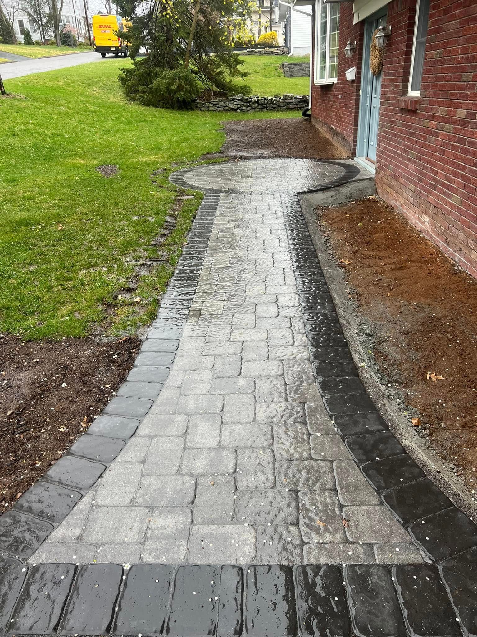 Brick pathway leading to a red brick building, wet from rain. Green grass on the left, dark border around pathway.