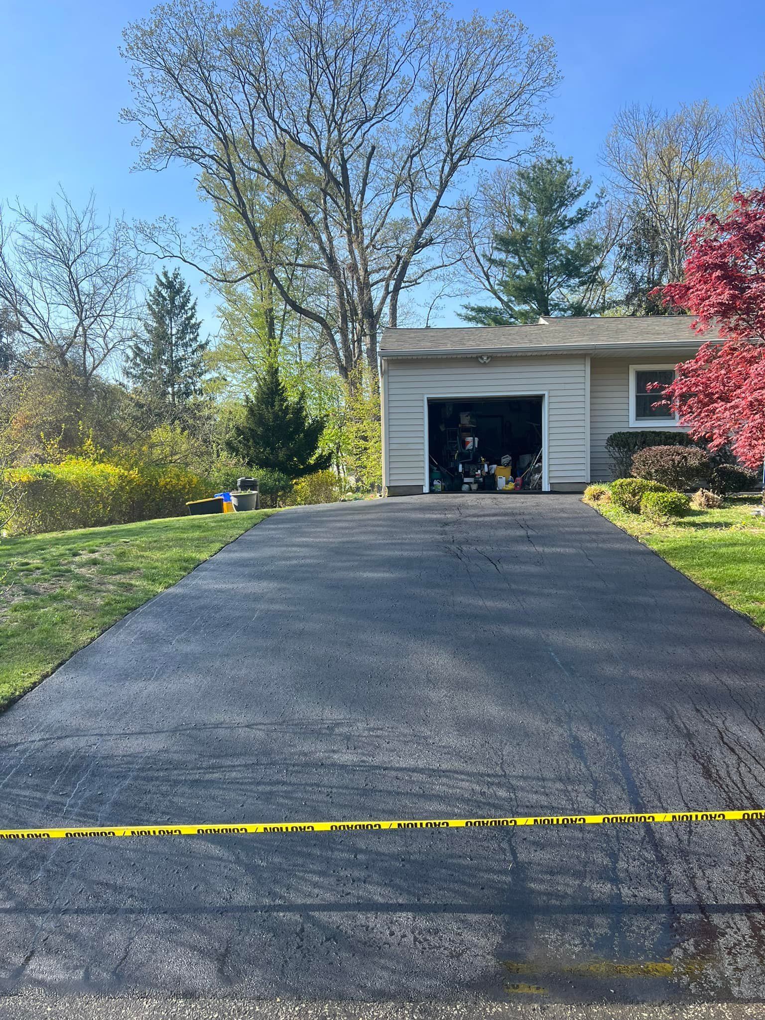 Paved driveway leading to a garage. Yellow caution tape across the bottom. Trees and a blue sky in the background.