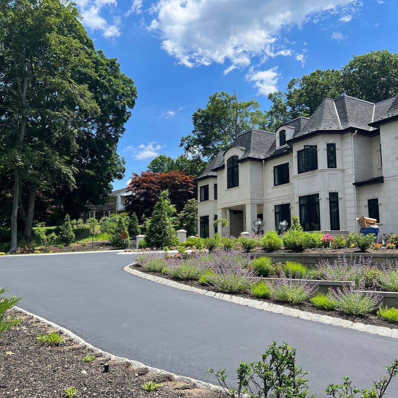 A large, light-colored house with dark roof set on a paved driveway surrounded by lush greenery and flowers under a blue sky.