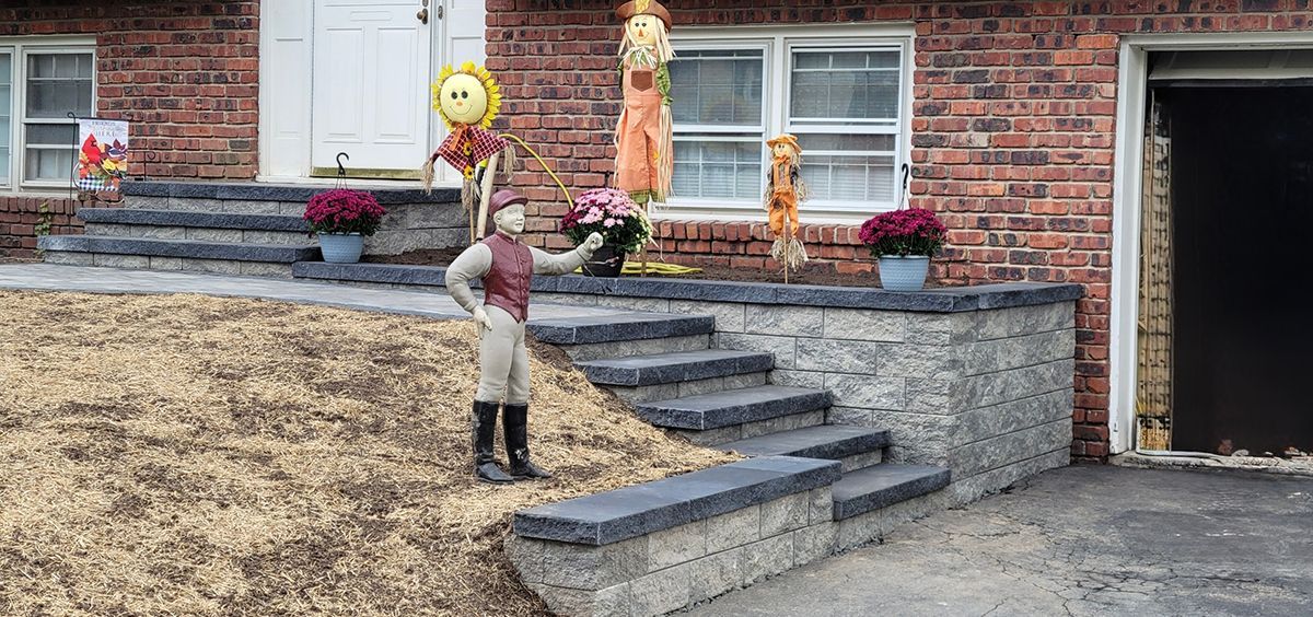 A figurine in riding attire stands on steps decorated for fall, with a brick house in the background.