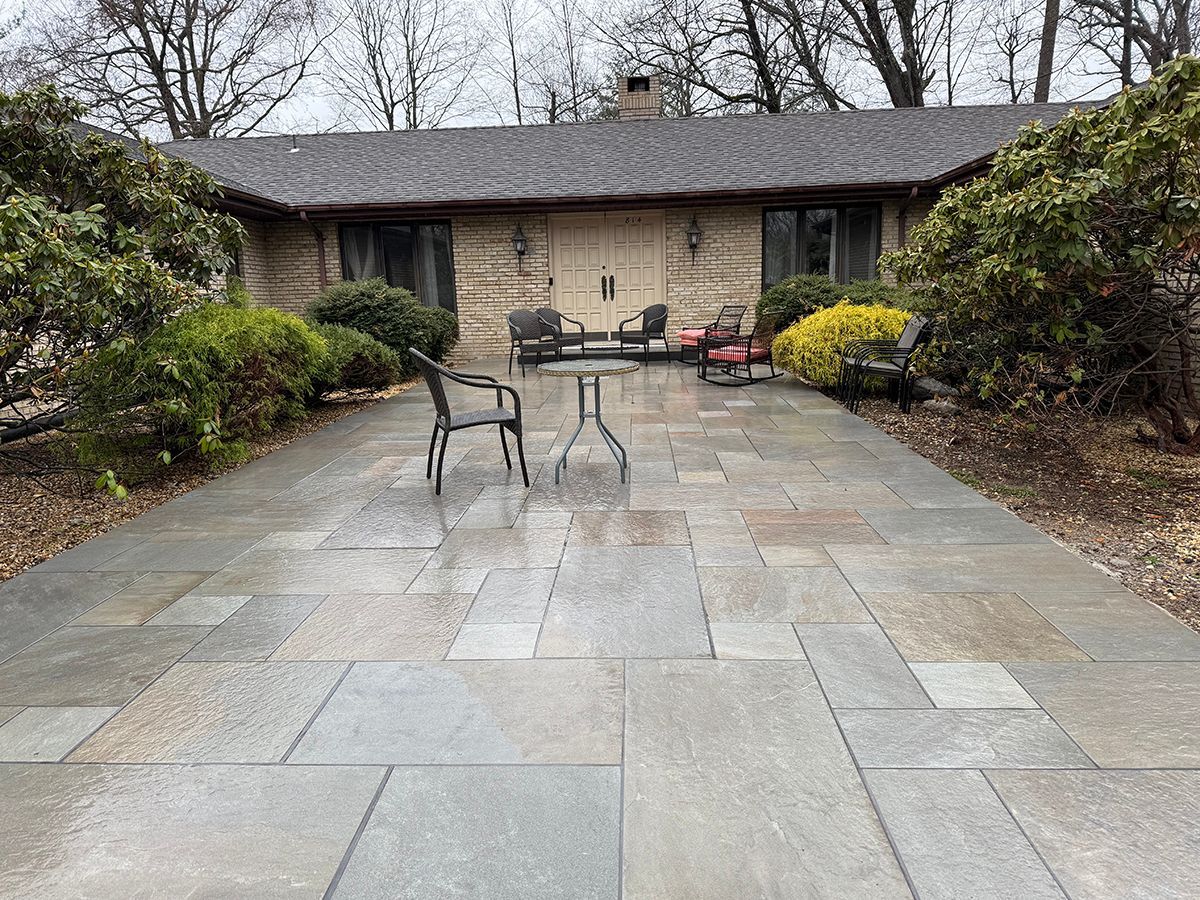 Stone patio in front of a tan brick house with trees and bushes. Outdoor seating visible.