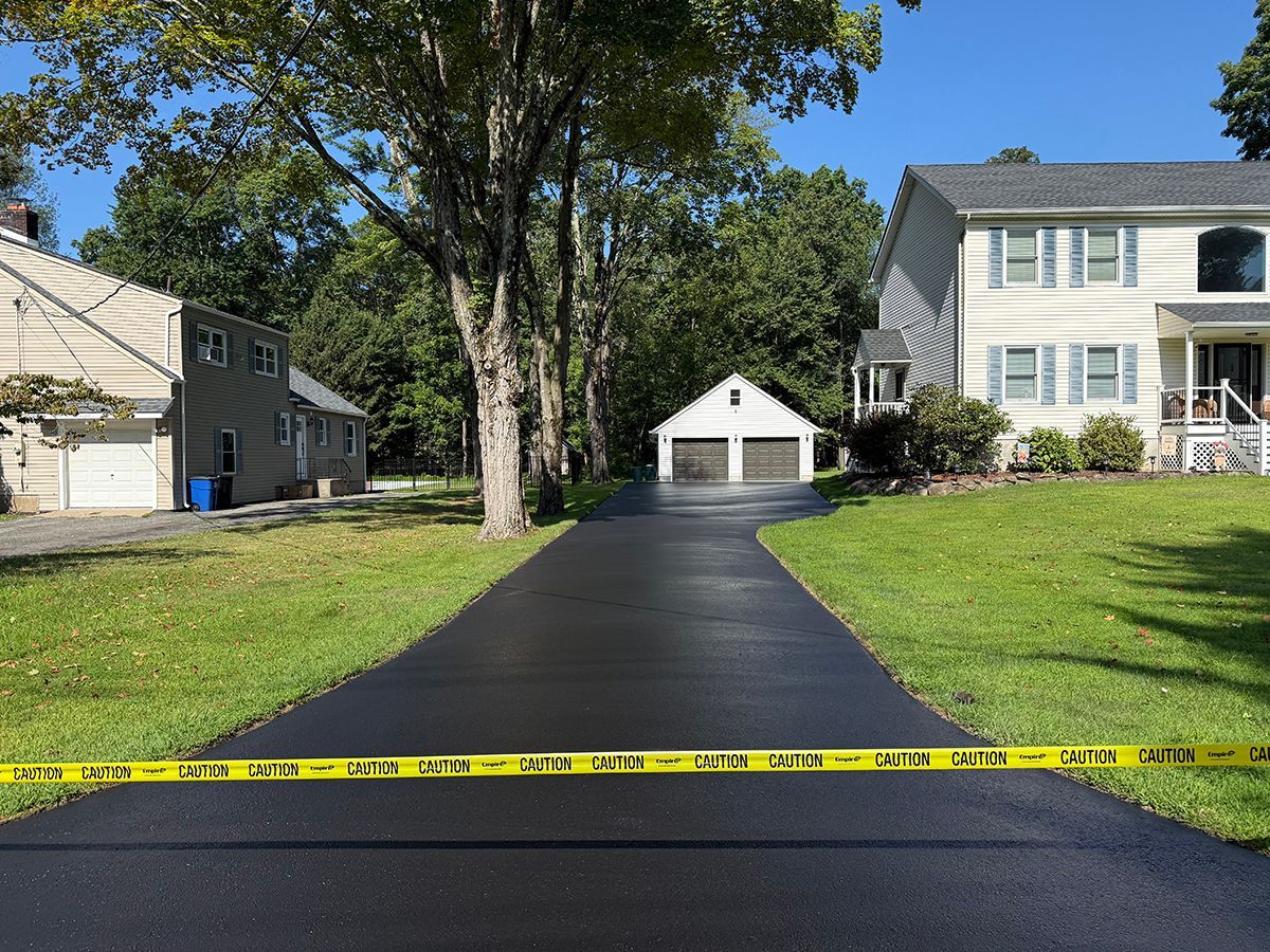 Newly paved black driveway with caution tape, leading to a two-car garage between two houses, sunny day.