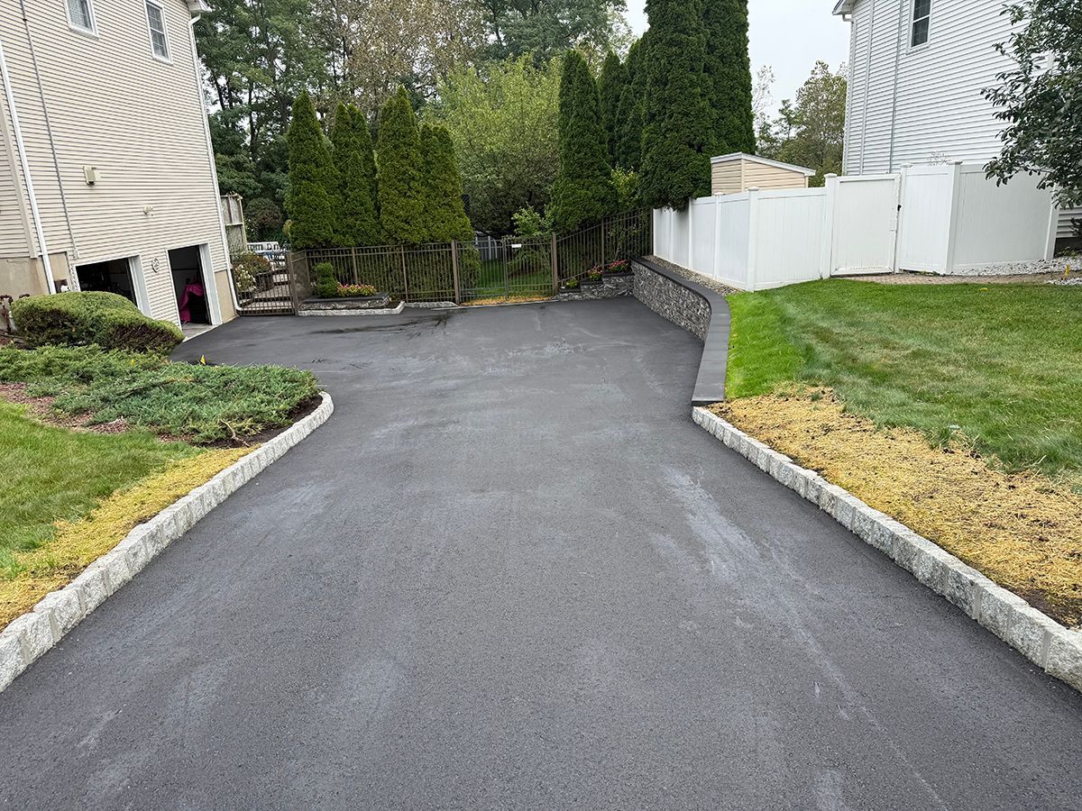 Paved driveway with granite borders, leading to a yard with shrubs, trees, and a white fence.