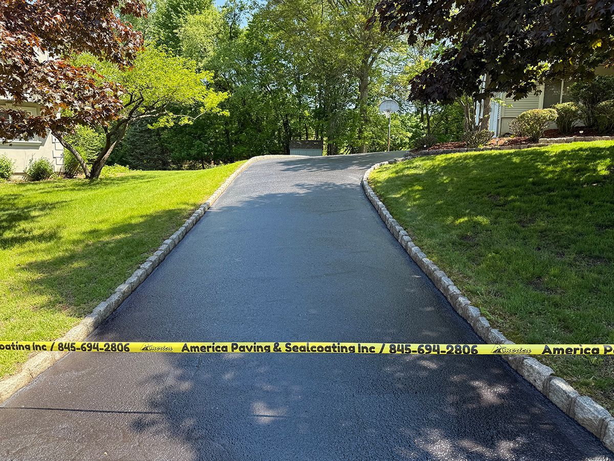 Freshly paved asphalt driveway blocked by yellow caution tape, flanked by green grass and trees.