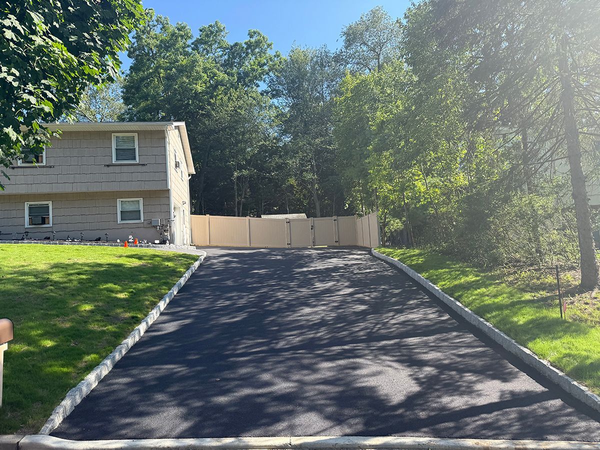 Asphalt driveway leading to a light-colored fence and house. Green grass on both sides, trees in the background.