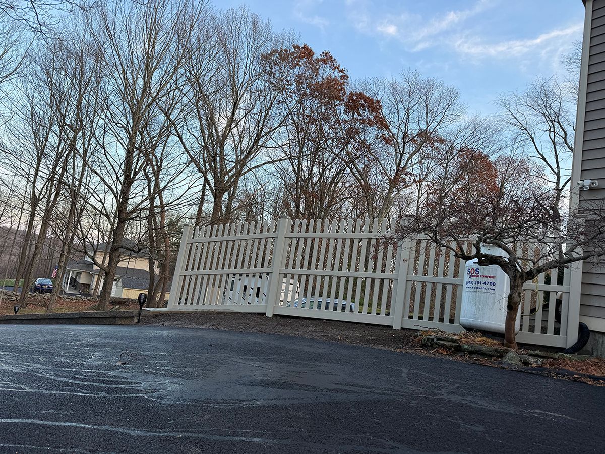 White picket fence in front of bare trees and blue sky. Black asphalt driveway.