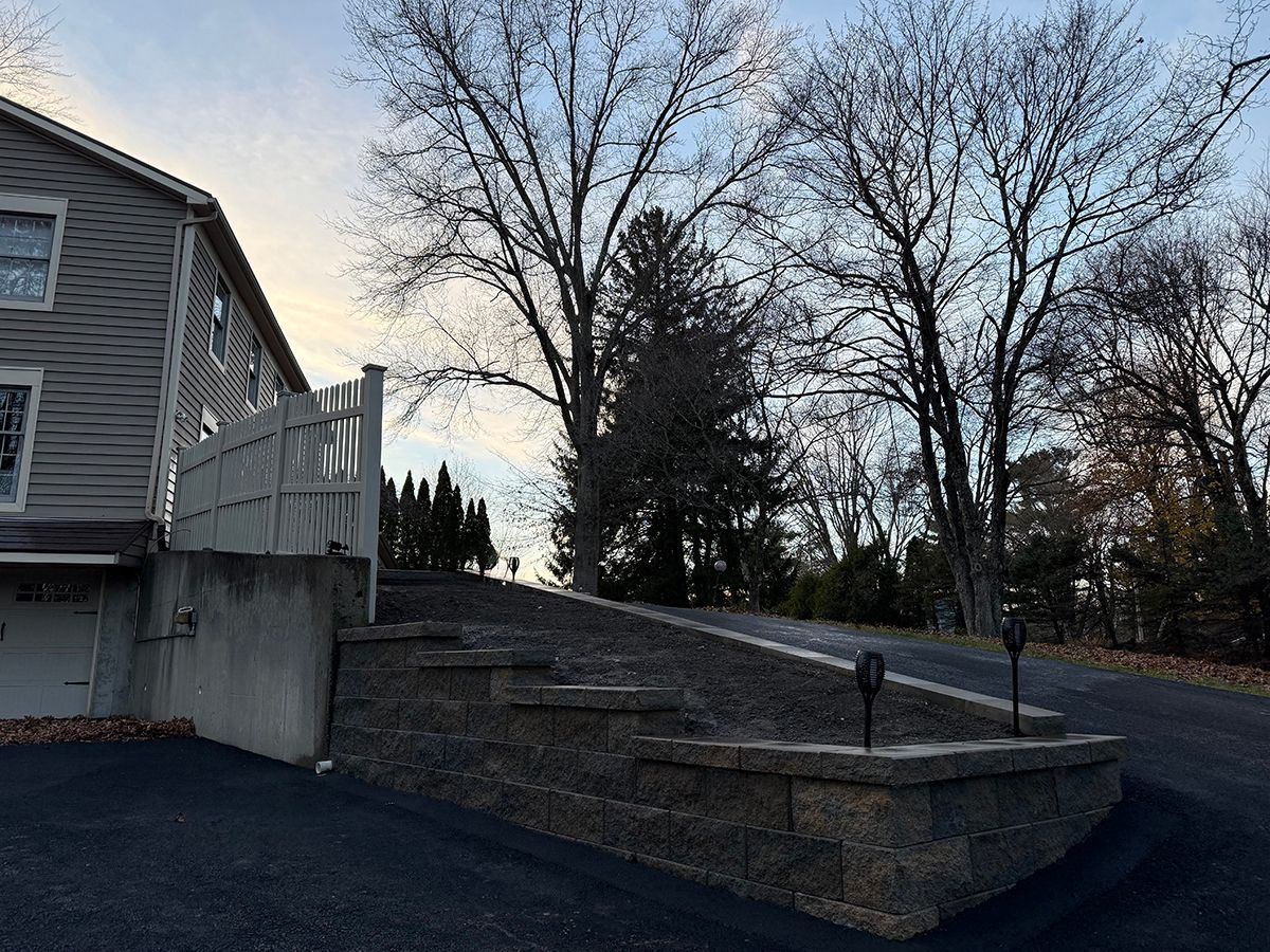 House with a dark asphalt driveway, stone steps, and a white fence in front of bare trees.