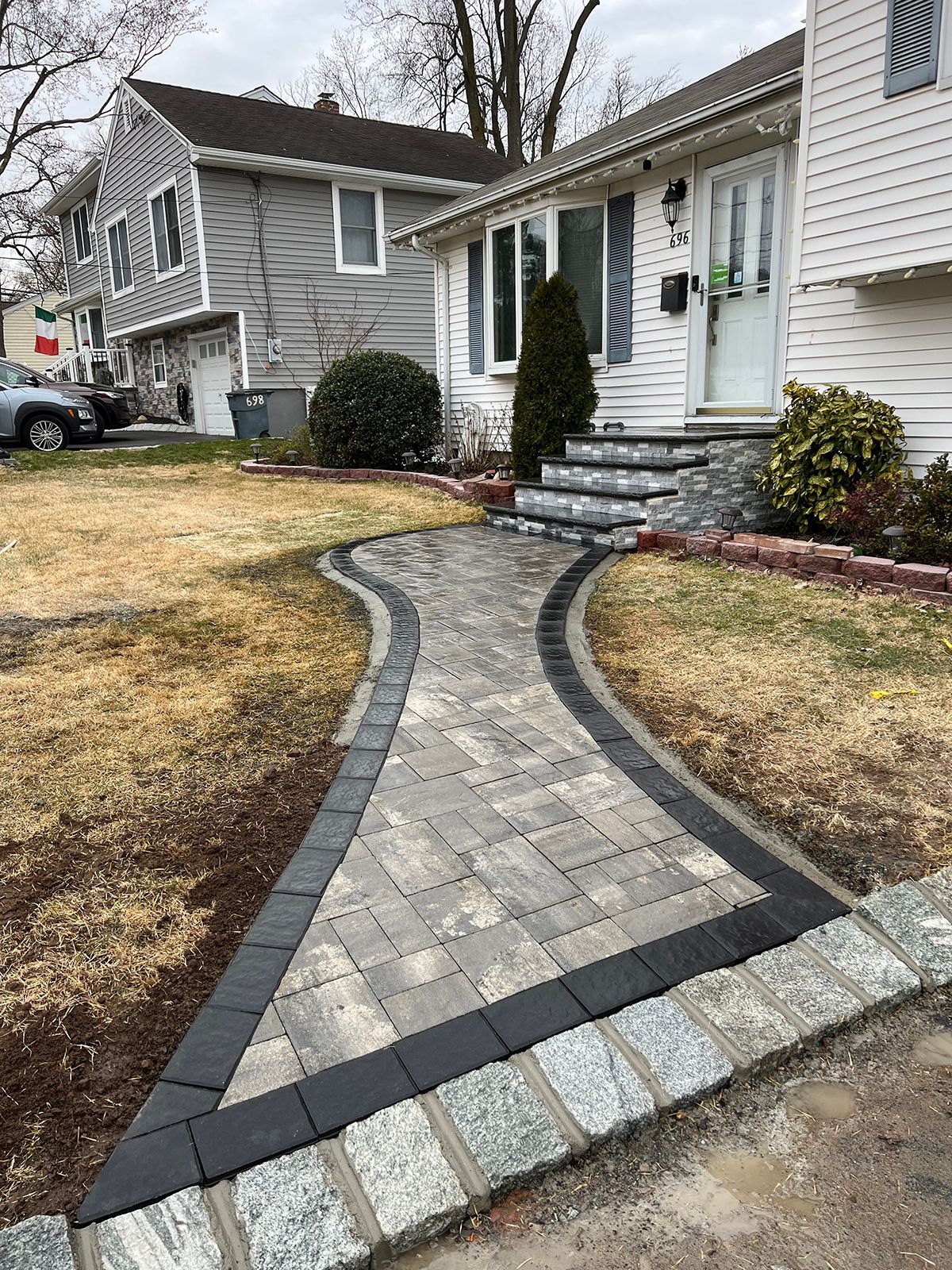 Brick pathway leading to a house with front steps, flanked by brown grass and other houses.