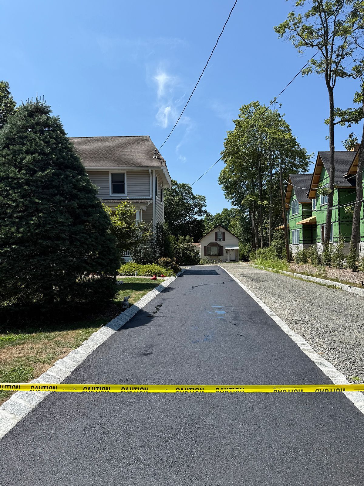 Asphalt driveway with caution tape, houses on either side, blue sky.