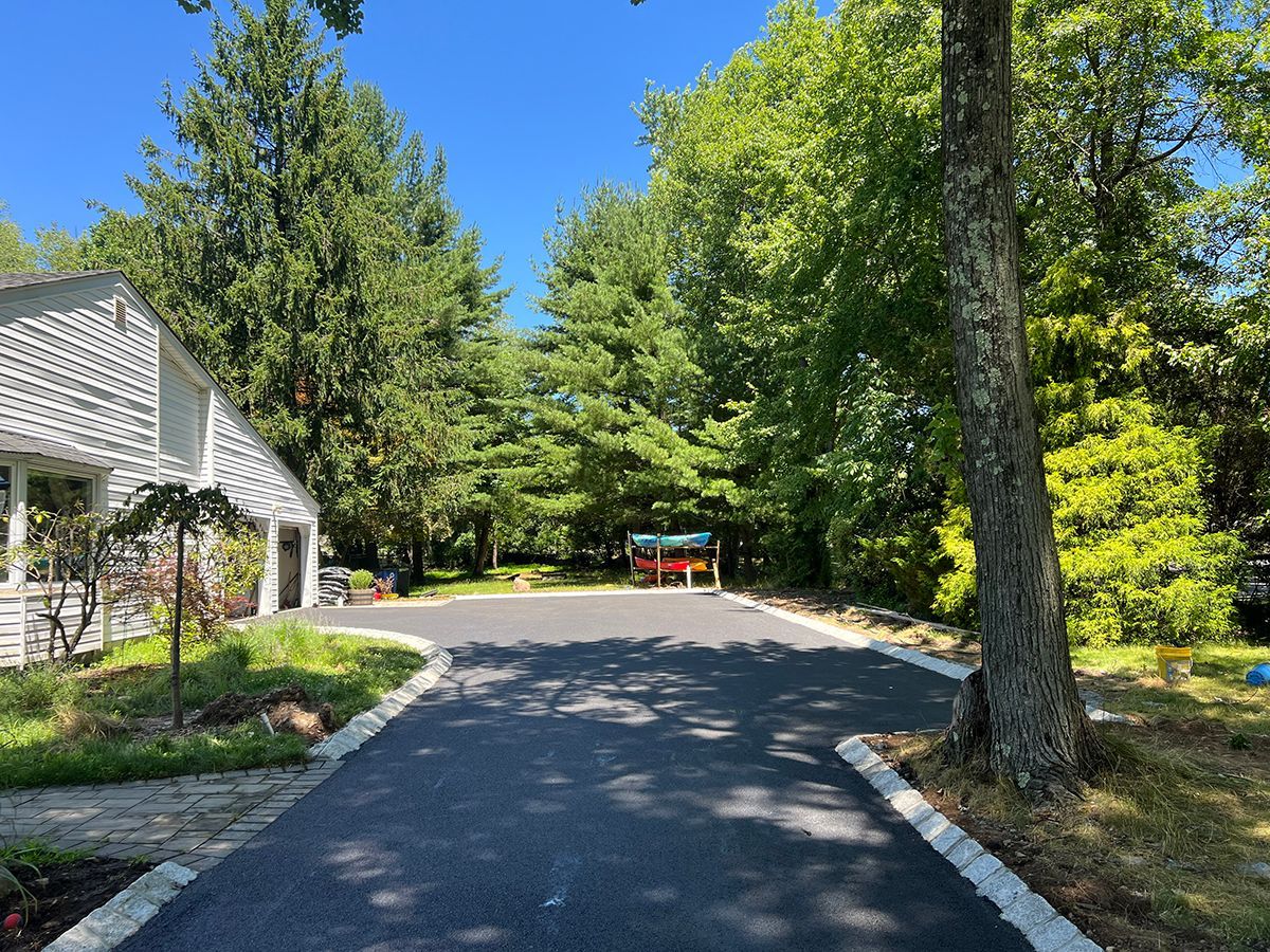 Asphalt driveway leading towards trees, bordered by curb stones and a lawn. A house is on the left. Bright sunny day.