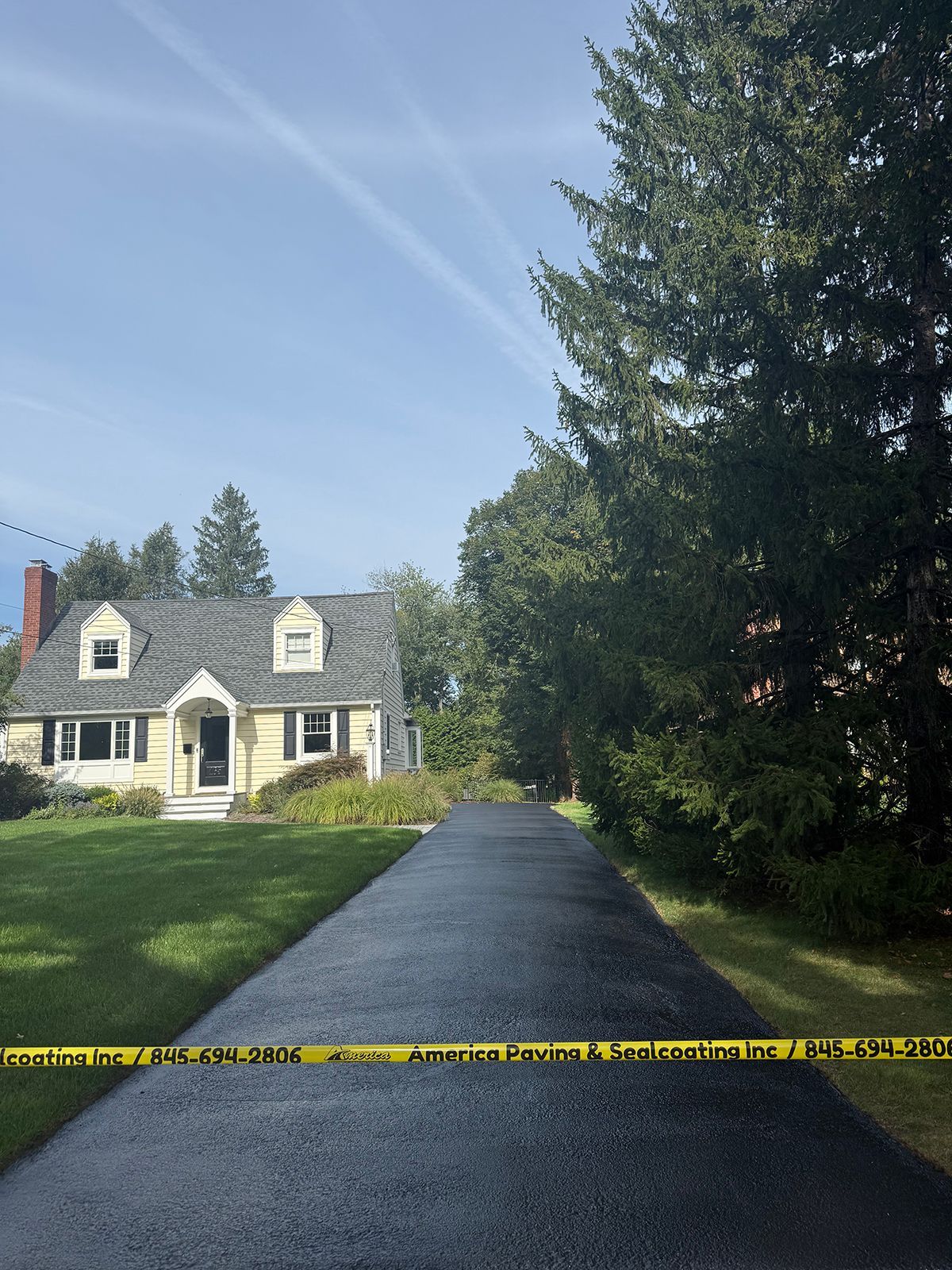 Asphalt driveway leading to a yellow house with a green lawn. Yellow caution tape is across the driveway.
