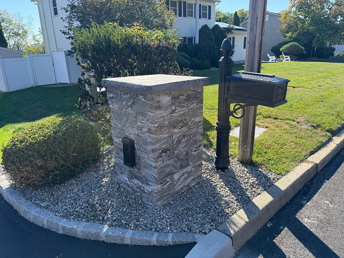 Stone mailbox post with a black mailbox, next to a grassy lawn and a house.