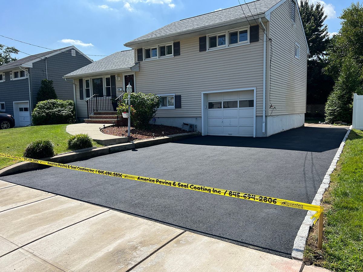 Newly paved black asphalt driveway in front of a two-story beige house with a garage; caution tape surrounds.