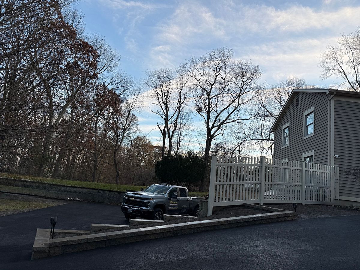 Grey pickup truck parked next to a house with white fence and trees under a cloudy sky.