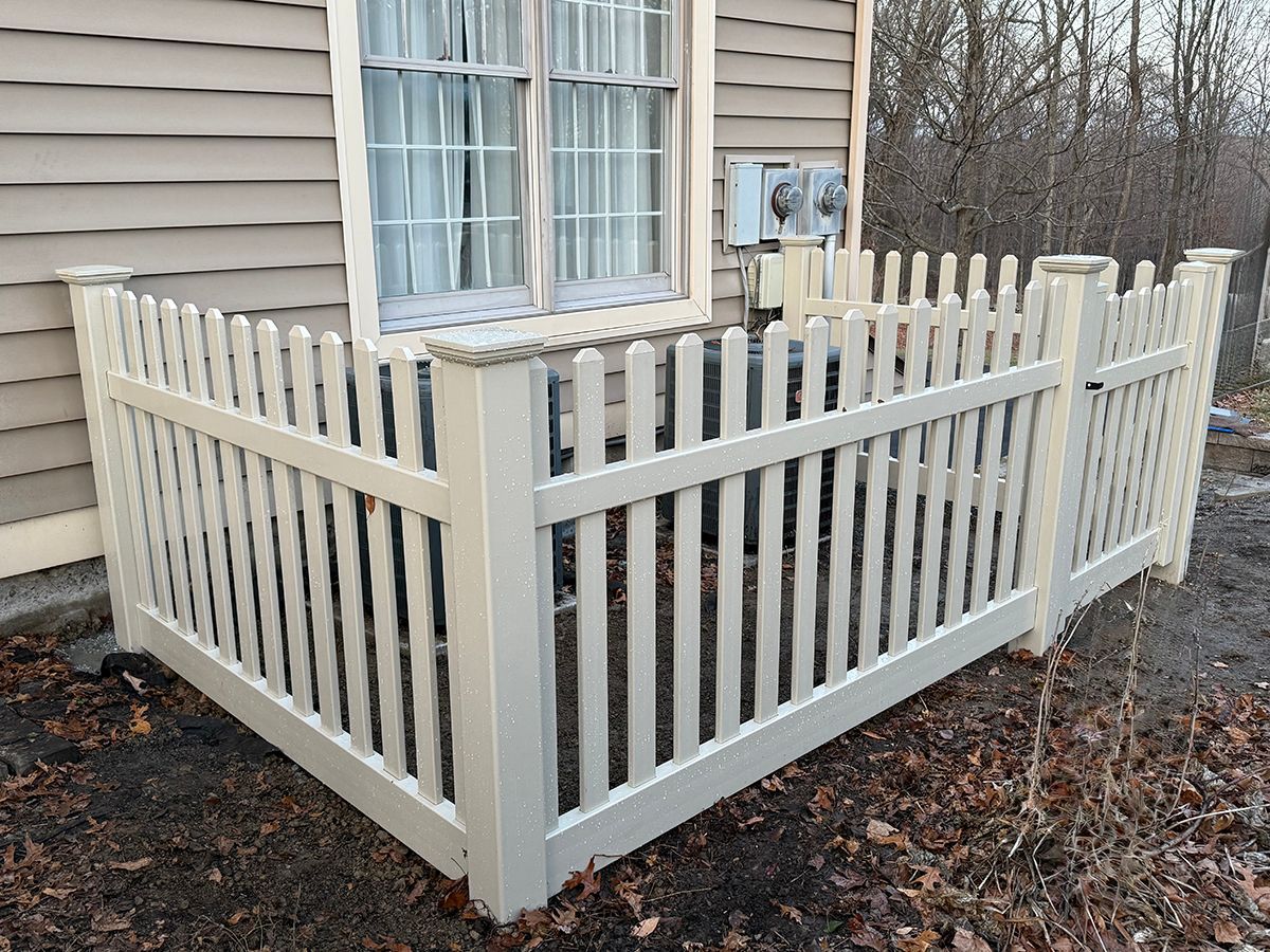 White picket fence surrounding an air conditioning unit outside a beige house window.