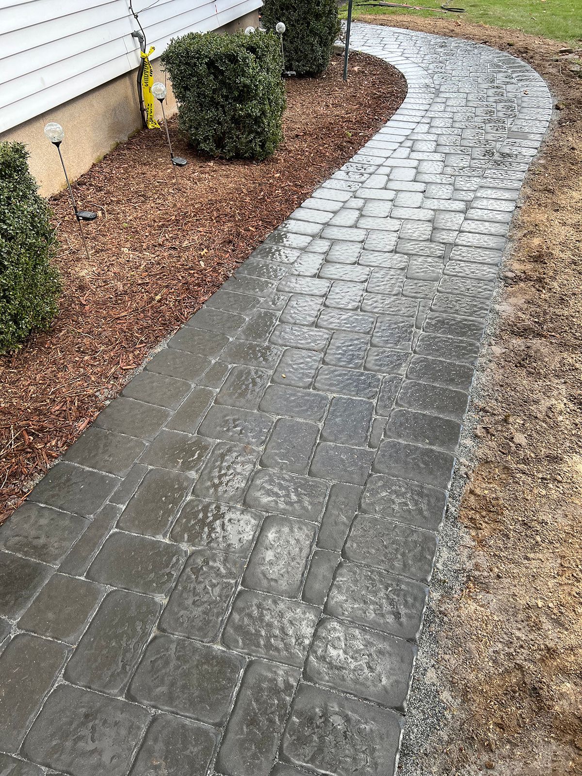 Brick pathway curves alongside a building, bordered by mulch and shrubs.