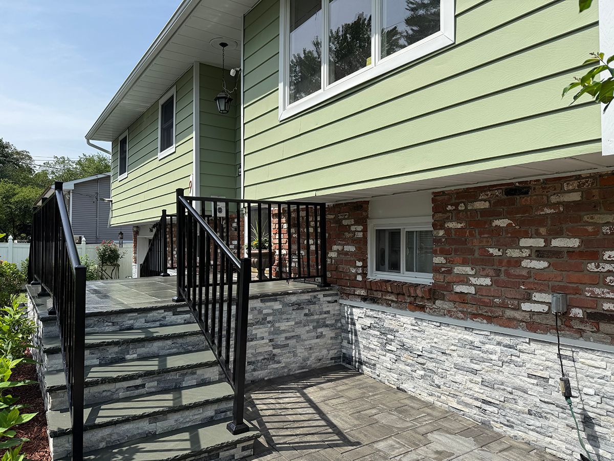 Two-story house with green siding, brick foundation, steps with black railing, and stone facade.
