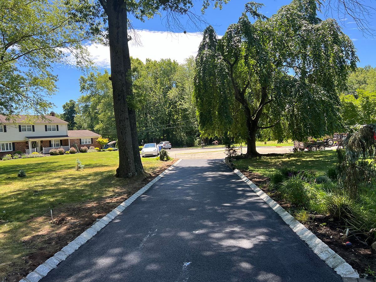 Asphalt driveway lined with stone, trees, and houses on a sunny day.