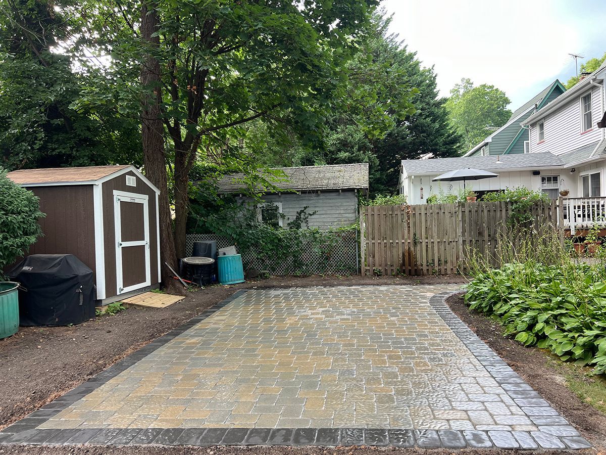 Patio with brick pavers, surrounded by plants and a fence, with a shed in the background.