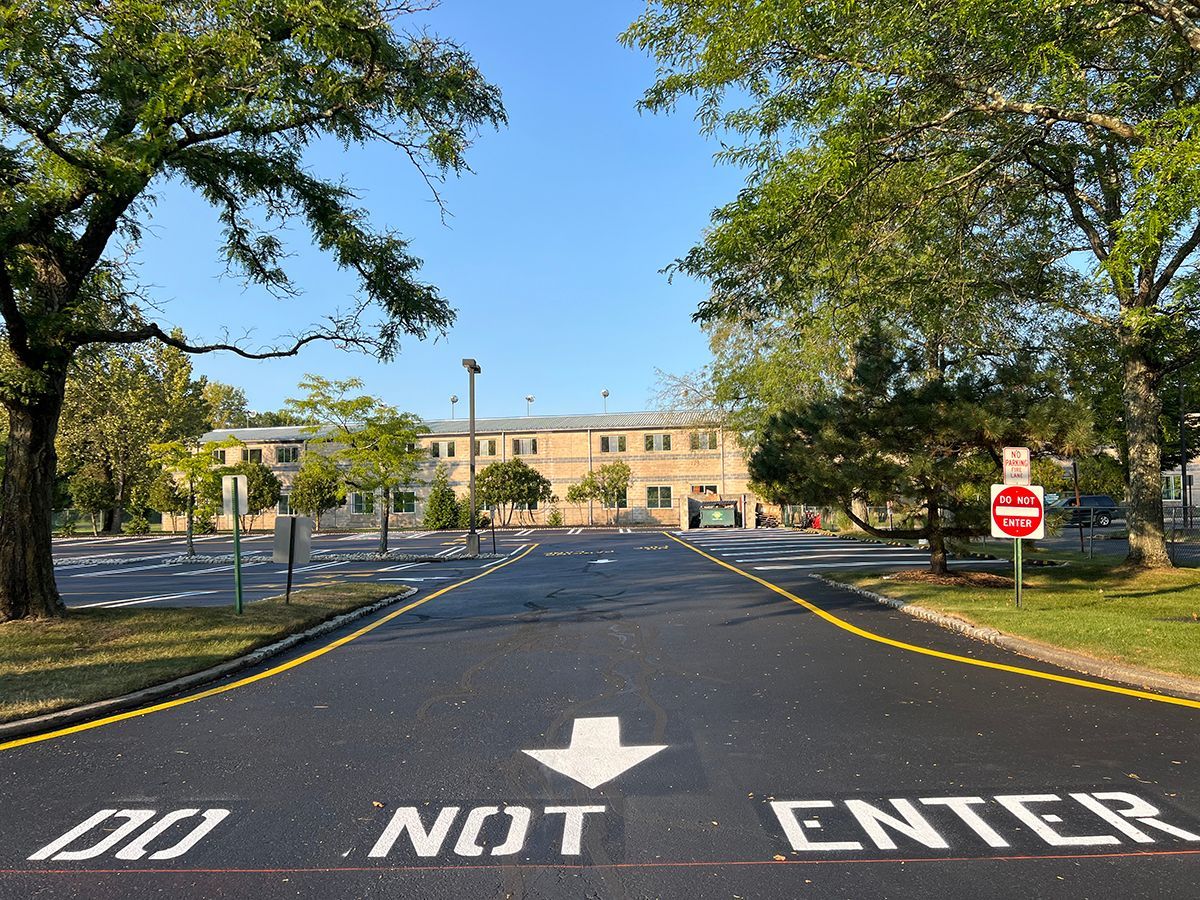 Paved road with marking leads to a beige building, flanked by trees under a blue sky.