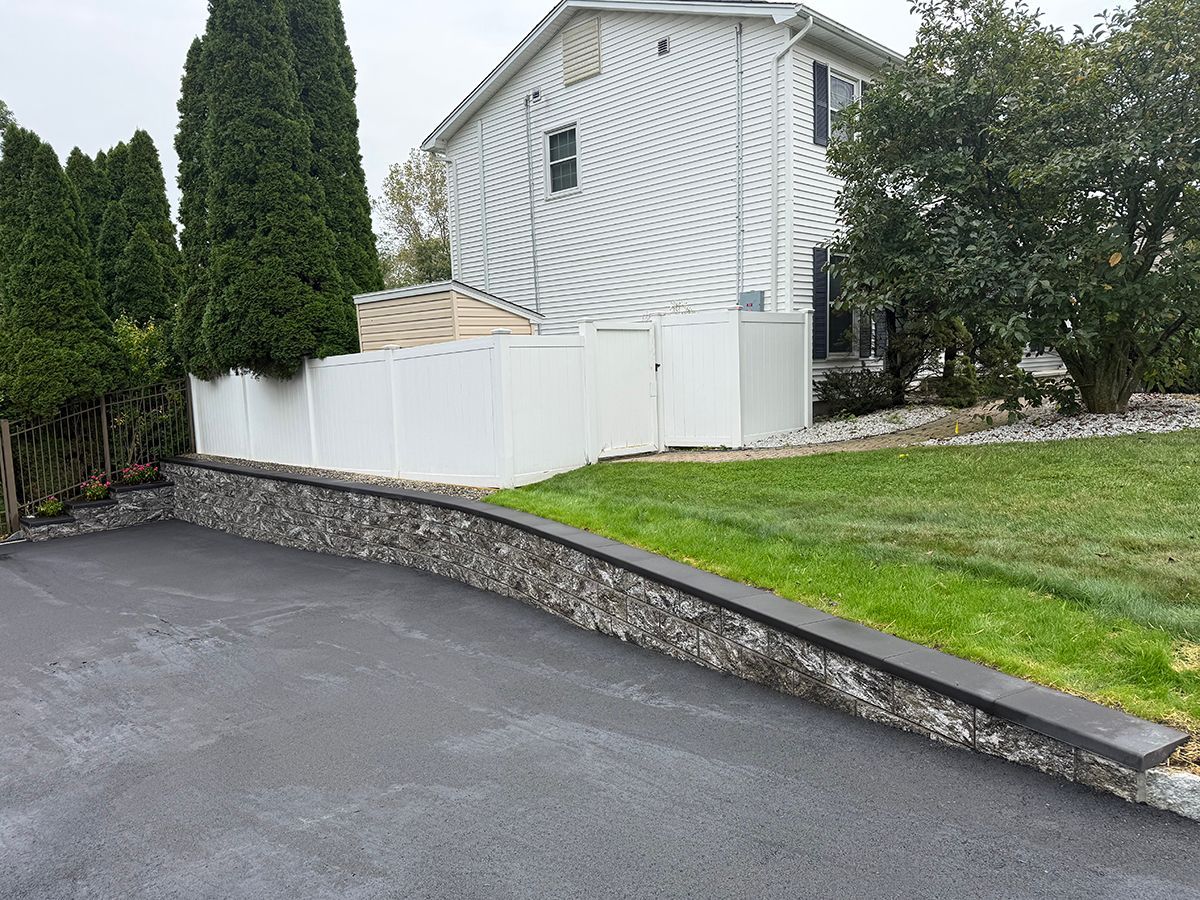 Asphalt driveway with stone retaining wall, white fence, and house with green lawn.