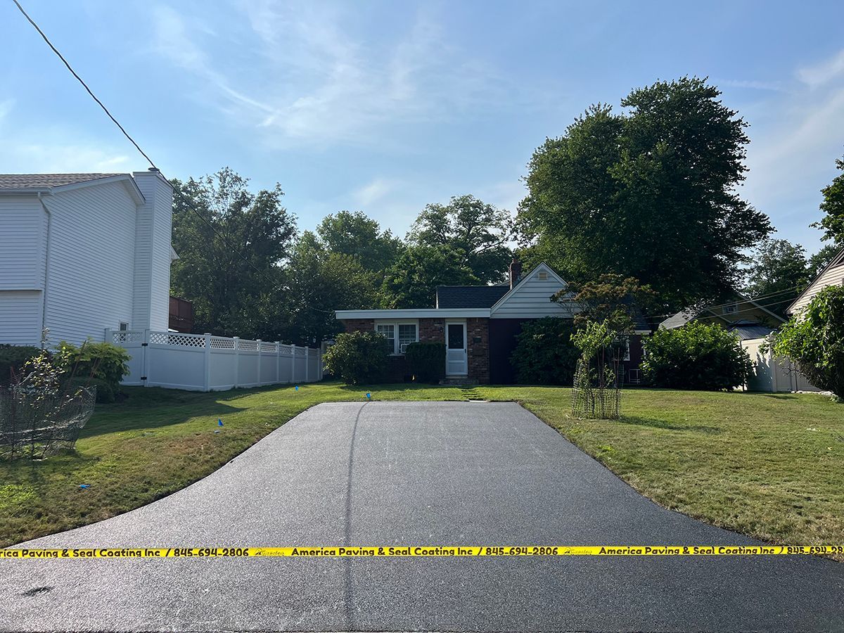 Asphalt driveway leading to a small house with a low roof, with trees and a blue sky in the background. Yellow caution tape is across the driveway.