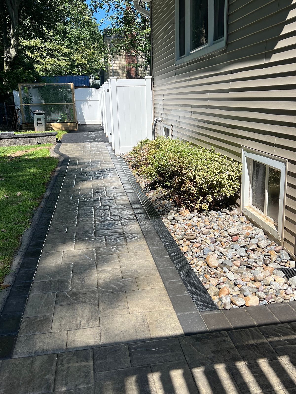 Paver pathway beside a house with a window, bushes, and a white fence.