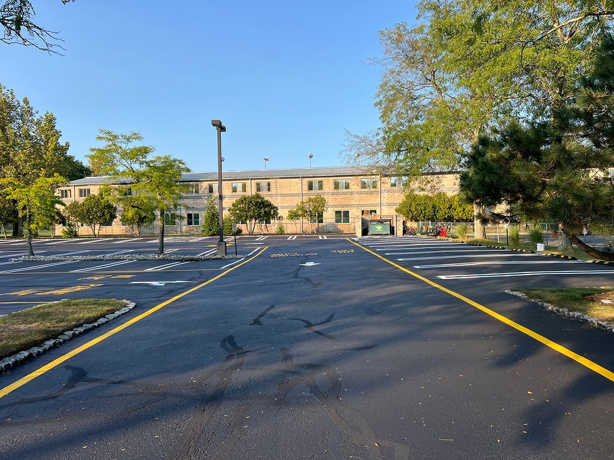 Asphalt parking lot with yellow lines, leading to a light-colored brick building. Trees frame the building.
