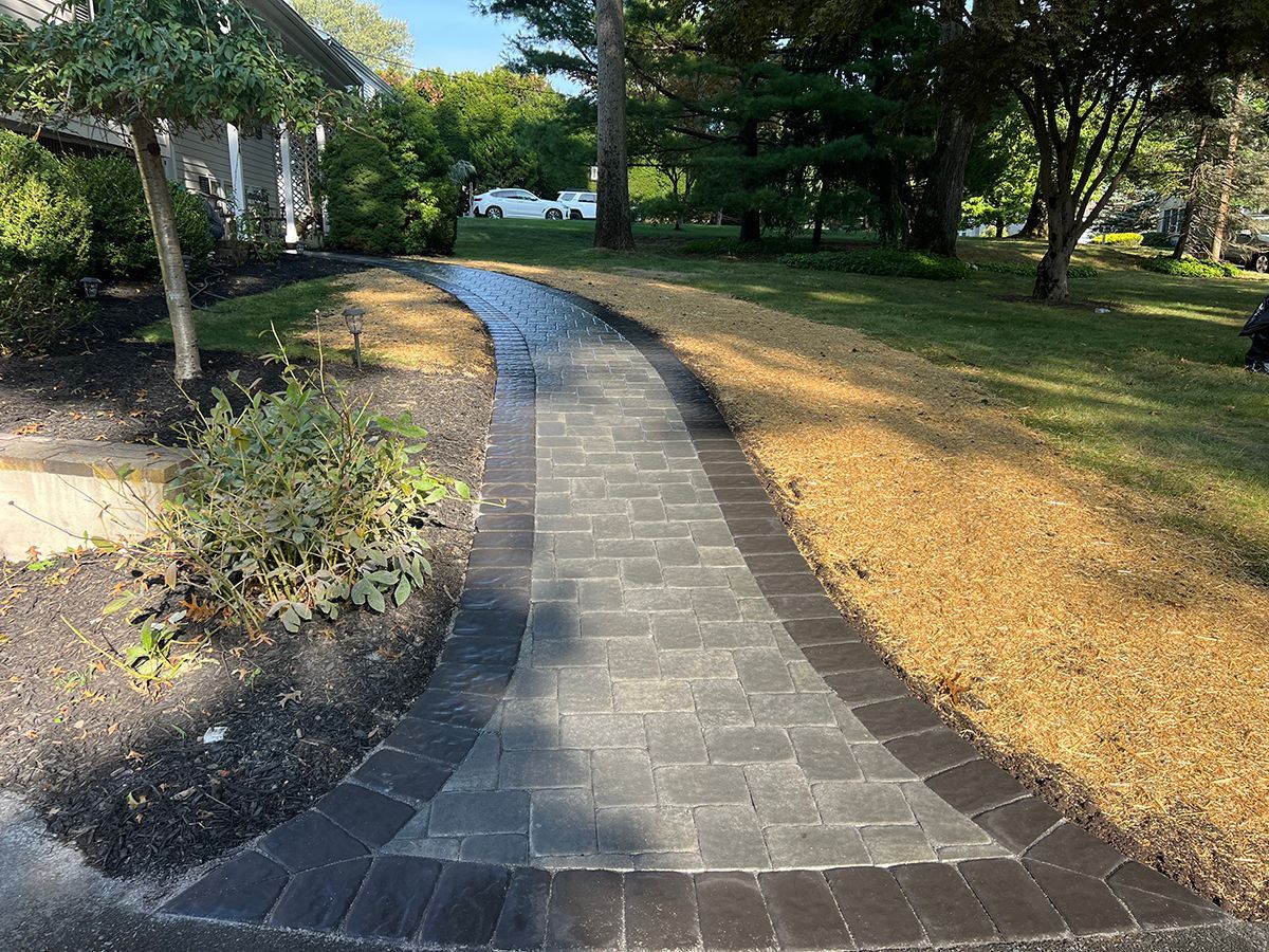 Brick pathway through a yard, bordered by dark stone, brown grass, and landscaping.