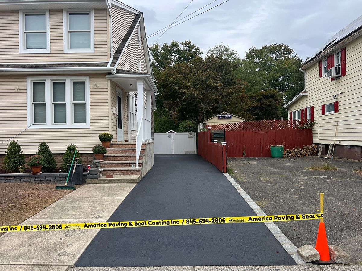 Freshly paved asphalt driveway blocked by caution tape; two-story house on left, red house on right.