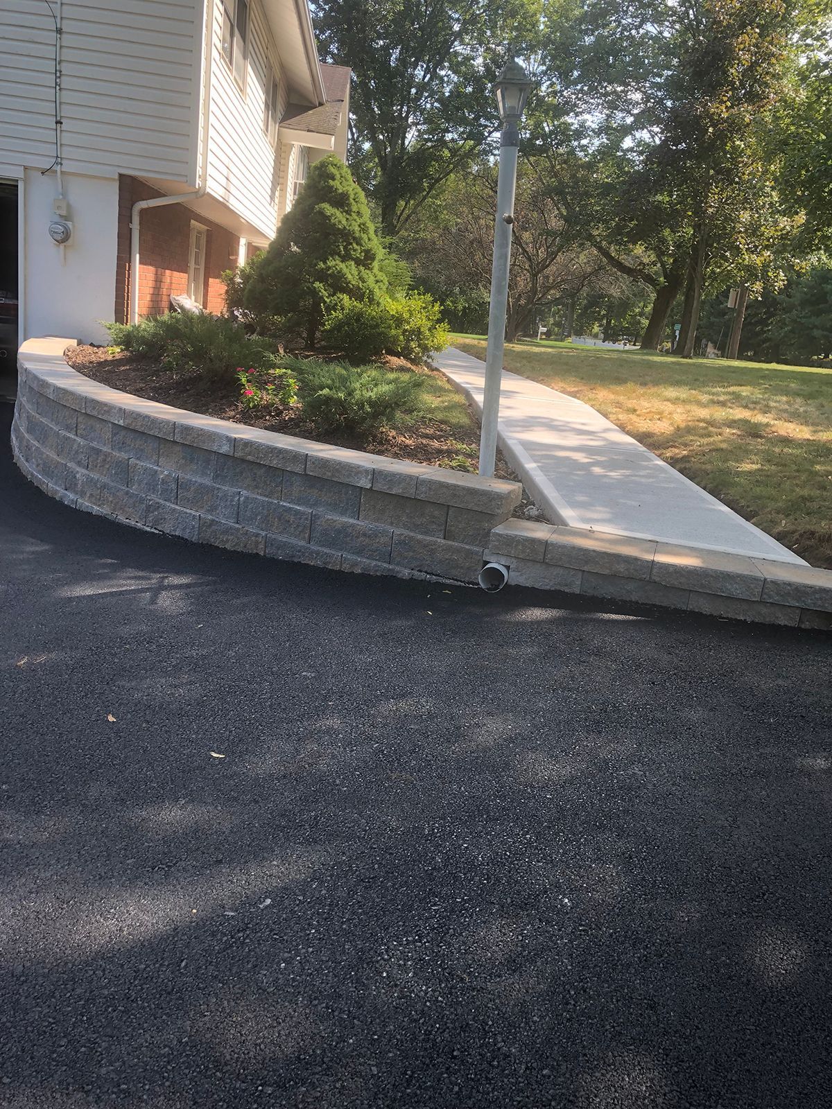 Brick retaining wall curves around a landscaped area next to a driveway and concrete sidewalk.