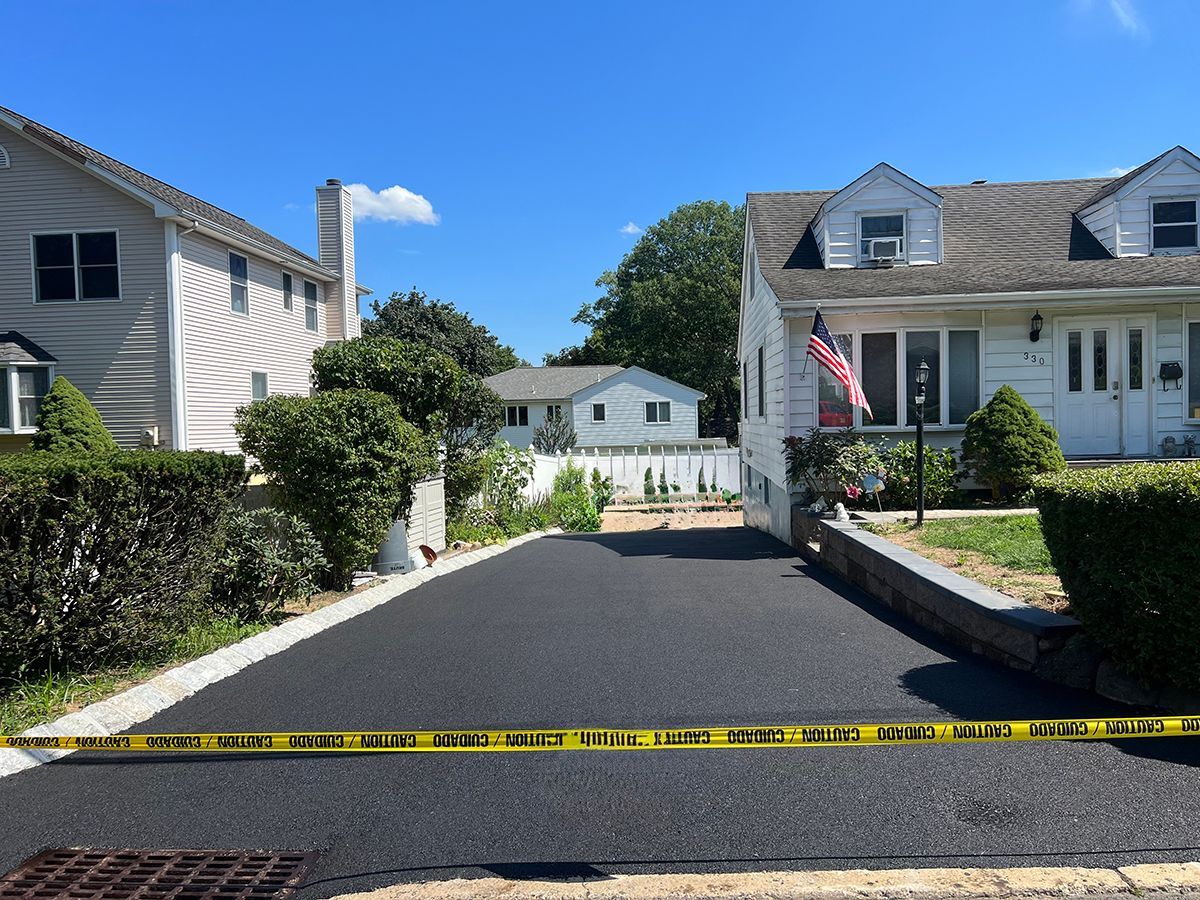 Newly paved driveway in a suburban neighborhood, with houses on either side and caution tape.
